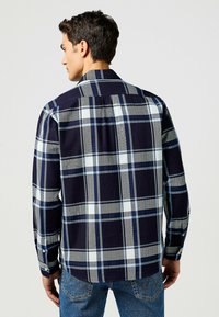Camicia a maniche lunghe a quadri in blu navy, grigio e azzurro. Presenta un taglio dritto e un dettaglio di yoke posteriore singolo.