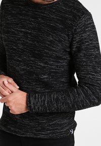 Pull noir à motif texturé en maille avec des fils gris. Il présente un col rond et des manches longues, ajustées aux poignets.