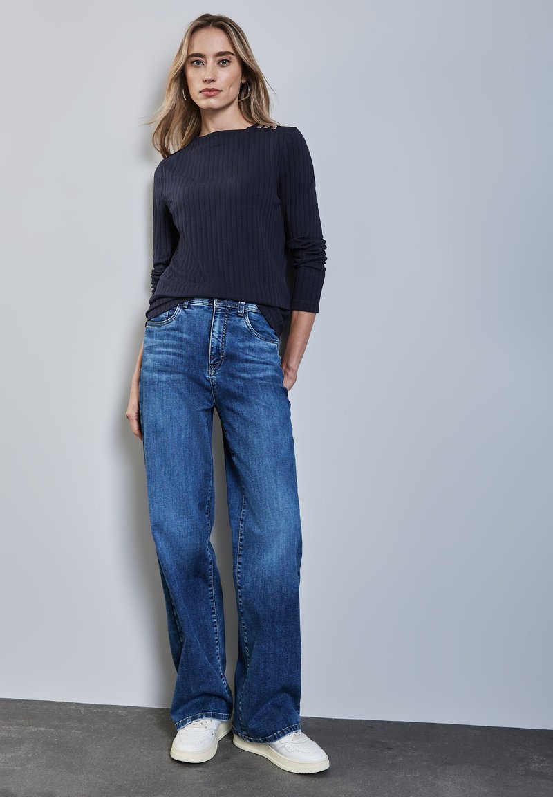 Maglietta a maniche lunghe a coste blu navy abbinata a jeans a gamba larga blu chiaro e sneakers bianche. Outfit semplice e casual su uno sfondo neutro.