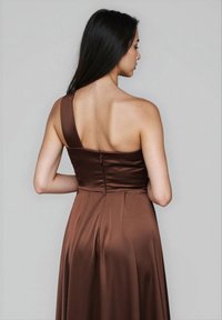 Donna con lunghi capelli scuri che indossa un abito in satin marrone a spalla unica, vista da dietro su uno sfondo grigio uniforme.