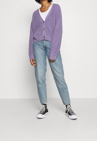 Lila stickad kofta med V-ringning, träknappar, kombinerad med ljusblå jeans och svarta högstövlade sneakers.