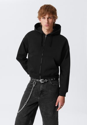 Sudadera negra con cremallera y bolsillo frontal, con cordón ajustable. Combinada con unos vaqueros gris oscuro que tienen un accesorio de cadena y un cinturón con tachuelas.