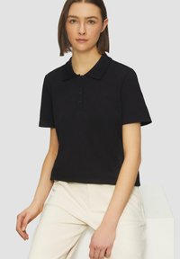 Polo noir à manches courtes, col classique et patte de boutonnage. Porté avec un pantalon couleur crème, avec une texture lisse.