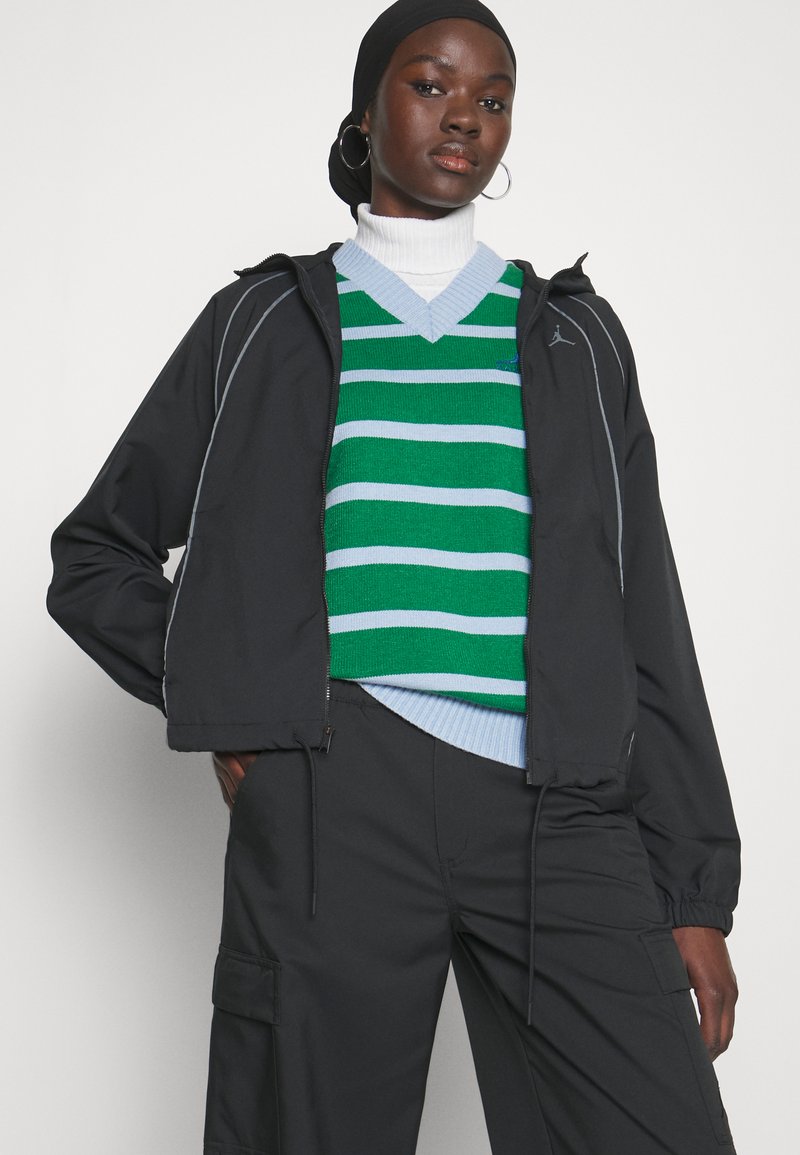 Chaqueta cortavientos negra con ribetes grises, superpuesta a un suéter a rayas verdes y azules. El modelo lleva pantalones cargo negros y un cuello de tortuga blanco.