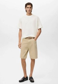 En ung mand står selvsikkert iført en hvid skjorte med korte ærmer, beige shorts og sorte loafers, foran en minimalistisk baggrund.