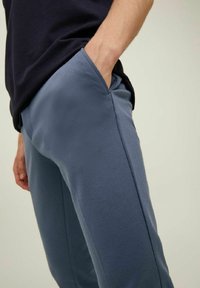 Persona vestida con pantalones azules, con una mano en el bolsillo y una camisa de un tono azul marino oscuro, de pie contra un fondo claro y sencillo.