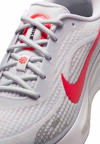 Scarpa da corsa Nike con tomaia in rete bianca, strisce grigie, accenti rosso vivo e lacci grigi. Presenta un logo distintivo e motivi texturizzati.