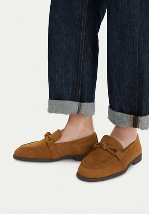 Brązowe mokasyny zamszowe z detalem węzła, noszone z ciemnymi, podwiniętymi dżinsami na jasnym tle.