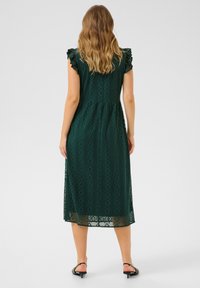 Grön spetsklänning med ett diamantmönster, korta volangärmar, figurnära midja och midi-längd, i kombination med svarta klacksandaler.