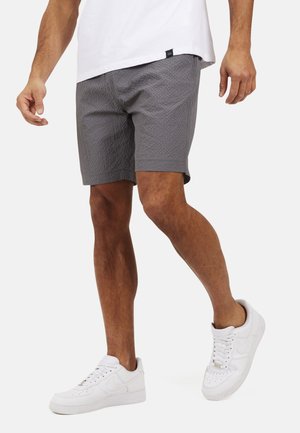 Uomo che indossa pantaloncini grigi con trama, scarpe da ginnastica bianche e una maglietta bianca, camminando con una gamba leggermente sollevata.