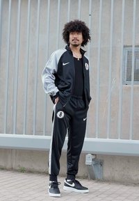 Μαύρο και γκρι σπορ σετ Nike με λογότυπο που περιλαμβάνει το "B", φορεμένο με μαύρη μπλούζα και μαύρα αθλητικά παπούτσια. Το ύφασμα φαίνεται λείο και αθλητικό.