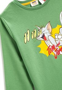 Sudadera de algodón verde que presenta un gráfico de un gato gris de dibujos animados con grandes ojos estilizados y el texto "Tom y Jerry" en rojo. Textura suave, cuello redondo.