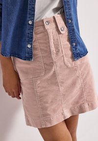 Kvinna iklädd en ljus rosa manchesterkjol med stora framfickor och en blå knappad denimskjorta över en vit topp.