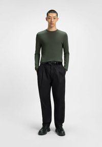 Langärmliges, olivgrünes Oberteil, kombiniert mit lockeren, gefalteten schwarzen Hosen. Ein schwarzer Gürtel und schwarze Schuhe runden das Outfit ab. Neutraler Hintergrund.