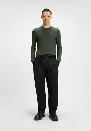 Long-sleeve, olive green top paired with loose, pleated black pants. Black belt and black shoes complete the outfit. Neutral background.