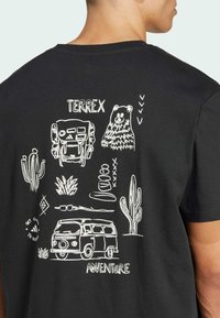 Black T-shirt featuring white graphic illustrations of mountains, a bear, cactus, and a van, with text "TERREX ADVENTURE" in a casual font.