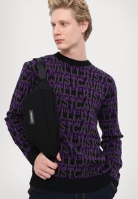 Purple and black sweater featuring repeating text design. Model wears a black crossbody bag with a zip and fabric strap.