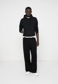 Hoodie en coton noir avec poche frontale, arborant un logo en relief, superposé sur un t-shirt blanc. Associé à un pantalon noir ample et des chaussures blanches.