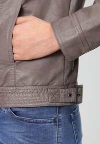 Chaqueta de cuero gris con una textura suave, que presenta un bolsillo lateral, detalles cosidos y una hebilla de cinturón en la cintura. Estilo casual mostrado.