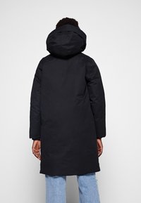 Parka negra con capucha, con una textura suave y mate. Mangas largas y un ajuste ligeramente holgado, llevada sobre unos vaqueros azul claro.