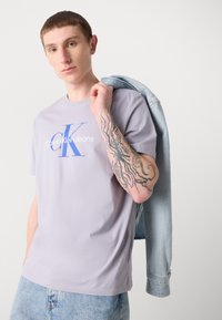 Light purple cotton t-shirt with blue text "Calvin Klein Jeans" and logo. Model holds a light denim jacket over shoulder.