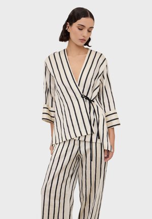 Kvinde iført cremefarvet omslagstop og vidde bukser med lodrette sorte striber, stående mod en enkel lys grå baggrund.