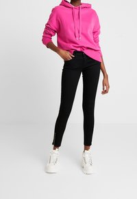 Sudadera rosa con cordones, jeans skinny negros con cremalleras en el dobladillo y zapatillas altas blancas. Fondo plano y neutro.