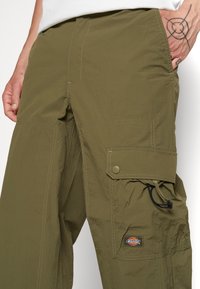 Man wearing olive green cargo pants with a flap pocket and black adjustable cord on the right thigh, showing a hand tattoo.