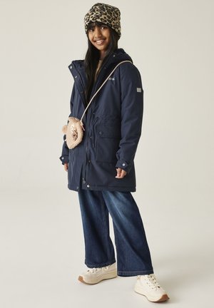 Uśmiechnięta młoda kobieta w granatowej parce, szerokich dżinsach, białych tenisówkach, kapeluszu w panterkę i małej torbie na ramię w kształcie misia.