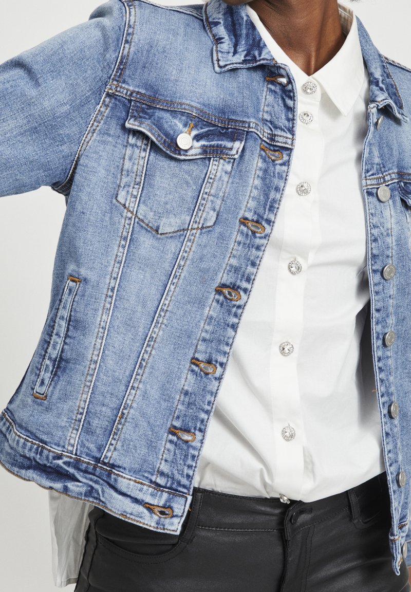 Personne portant une veste en jean bleu clair par-dessus une chemise blanche boutonnée et un pantalon noir, cadrée au niveau du cou et des hanches sur un fond uni.