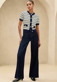 Cardigan a strisce navy e crema con maniche corte, chiusura a bottoni e due tasche frontali, abbinato a jeans a gamba larga in denim scuro e tacchi neri.