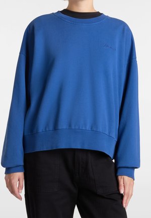 Persona con sudadera azul de manga larga con puños y dobladillo acanalados, combinada con pantalones negros de cintura alta, sobre un fondo liso.
