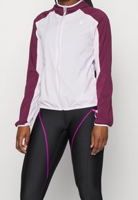 Veste légère à fermeture éclair blanche avec des accents bordeaux, dotée d'une capuche et de poignets élastiques, associée à un legging noir avec des détails roses.