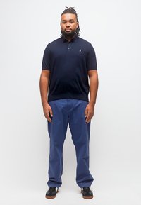 Άνδρας με dreadlocks φορώντας μαύρο πόλο πουκάμισο, μπλε παντελόνια και μαύρα αθλητικά παπούτσια, στέκεται μπροστά σε λευκό φόντο.