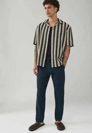 Camisa de manga corta con estampado en blanco y negro, pantalones azul marino y sandalias de cuero entrelazadas de color marrón oscuro. Presenta un corte holgado y un diseño de botones.