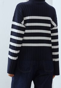 Pull en tricot côtelé présentant des rayures horizontales bleu marine et blanches, col montant et épaules tombantes, associé à un pantalon en denim foncé.