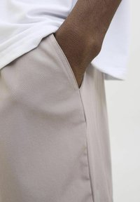 Pantalones gris claro con una textura suave, que presentan bolsillos laterales. Una camisa blanca es parcialmente visible por encima de la cintura, mostrando un ajuste relajado.