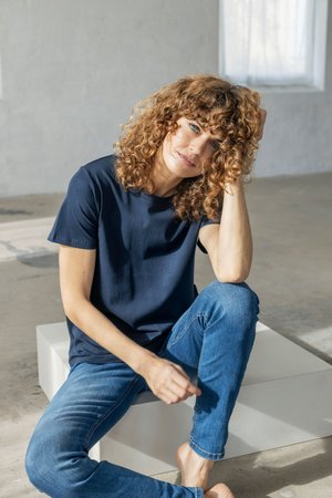 Woman with curly hair sitting barefoot on a white block indoors, wearing a navy t-shirt and blue jeans, resting her head on her hand.