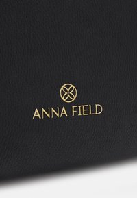 Borsa in ecopelle nera con texture presenta un logo "ANNA FIELD" in oro e un design con emblema floreale, enfatizzando un'estetica elegante e moderna.