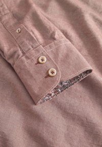 Cuff di camicia a righe marroni con due bottoni chiari e foderato con un motivo floreale. Il tessuto ha un aspetto strutturato.