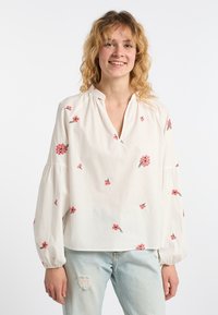 Weiße Bluse mit rosa floralem Stickerei, V-Ausschnitt und gerafften Details. Lange, luftige Ärmel mit elastischen Bündchen. Getragen mit hellblauen, beschädigten Jeans.