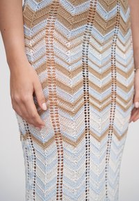 Mains reposant sur une robe tricotée avec des rayures chevron beige, bleues et blanches et des détails de points verticaux ouverts.