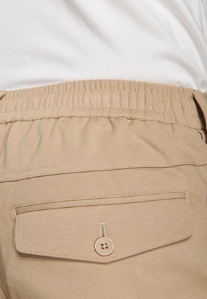 Lähikuva beigenvärisistä housuista, joissa on joustava vyötärö ja napillinen takatasku, yhdistettynä valkoiseen paitaan.