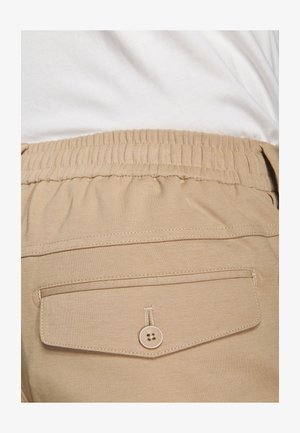 Lähikuva beigenvärisistä housuista, joissa on joustava vyötärö ja napillinen takatasku, yhdistettynä valkoiseen paitaan.