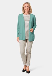 Helles türkisfarbenes Cardigan mit offener Vorderseite, langen Ärmeln und zwei Fronttaschen, getragen über einem hellen Satintop und beigen Hosen. Schwarzes Schuhwerk.