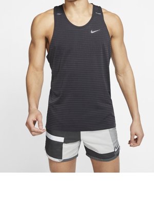 Personne musclée portant un débardeur noir sans manches Nike et un short Nike tricolore noir, blanc et gris, debout sur fond blanc.
