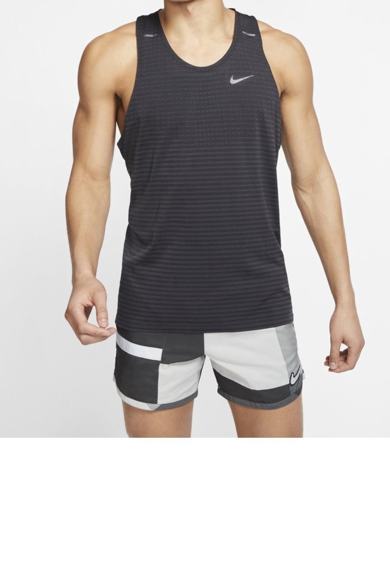 Personne musclée portant un débardeur noir sans manches Nike et un short Nike tricolore noir, blanc et gris, debout sur fond blanc.