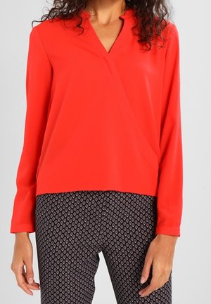 Rød langærmet bluse med V-udskæring og krydset design, lavet af et glat stof. Båret med mønstrede mørke bukser med små motiver.