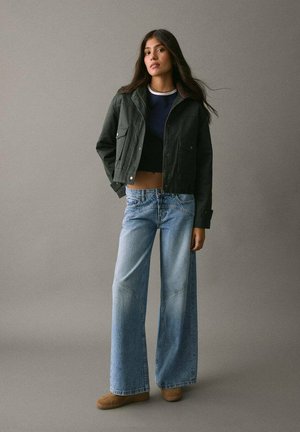 Donkergroene cropped jack met twee voorzakken, gecombineerd met lichtblauwe wijde jeans. Model staat voor een eenvoudige grijze achtergrond.