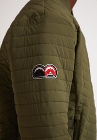 Giacca trapuntata leggera di colore verde oliva con un grande patch nero e rosso con motivi montani sulla manica sinistra.